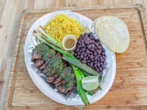 Churrasco on a white plate with black beans, vegetables and yellow rice on a wooden serving board at La Salsa Verde